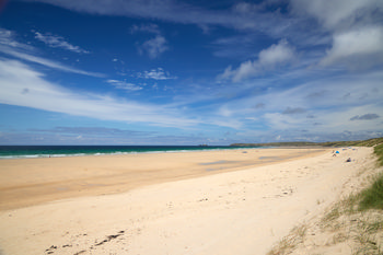 Upton Towans This landscape photograph depicts Upton Towans, a wide sandy beach along the coastline of Cornwall, England, in the United Kingdom. The scene shows the stretch of beach leading to the turquoise waters of the sea, with gentle waves visible in the distance. Captured in the early afternoon during the summer season, the image features blue skies with scattered clouds above the coast. The sandy dunes covered with grasses are visible in the foreground, adding natural detail to the coastal environment. In the distance, sections of the shoreline and headlands that are characteristic of Cornwall can be seen, reinforcing the beach setting in this part of England.